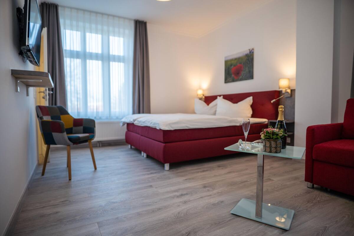 A hotel room with a bed, a TV, a red sofa, a glass coffee table, and a chair.
