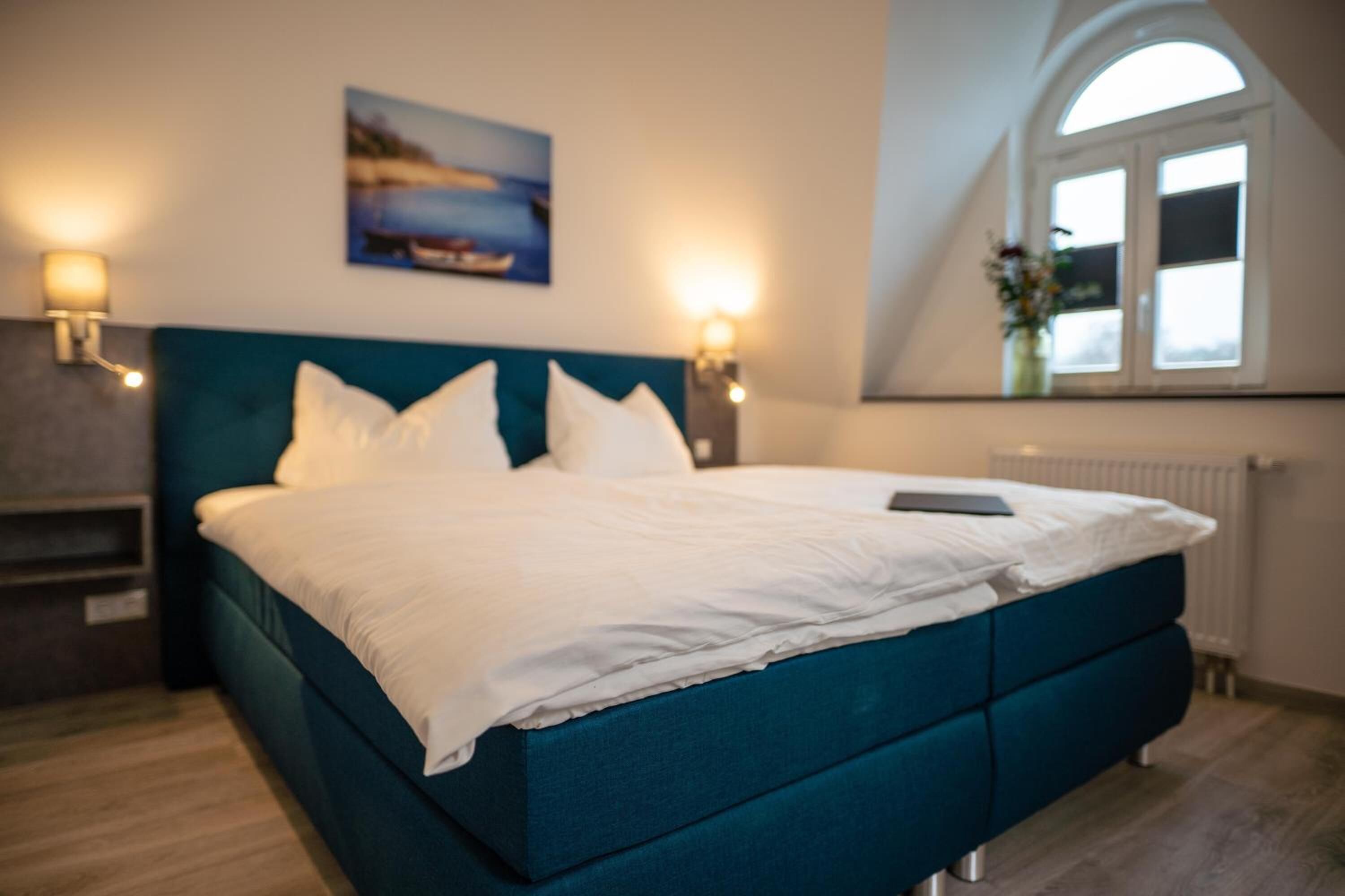 Une chambre à coucher avec un lit de couleur turquoise, du linge de lit blanc, deux lampes fixées au mur, un tableau au-dessus du lit et une fenêtre donnant sur un beau paysage.