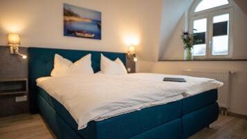 Une chambre à coucher avec un lit de couleur turquoise, du linge de lit blanc, deux lampes fixées au mur, un tableau au-dessus du lit et une fenêtre donnant sur un beau paysage.