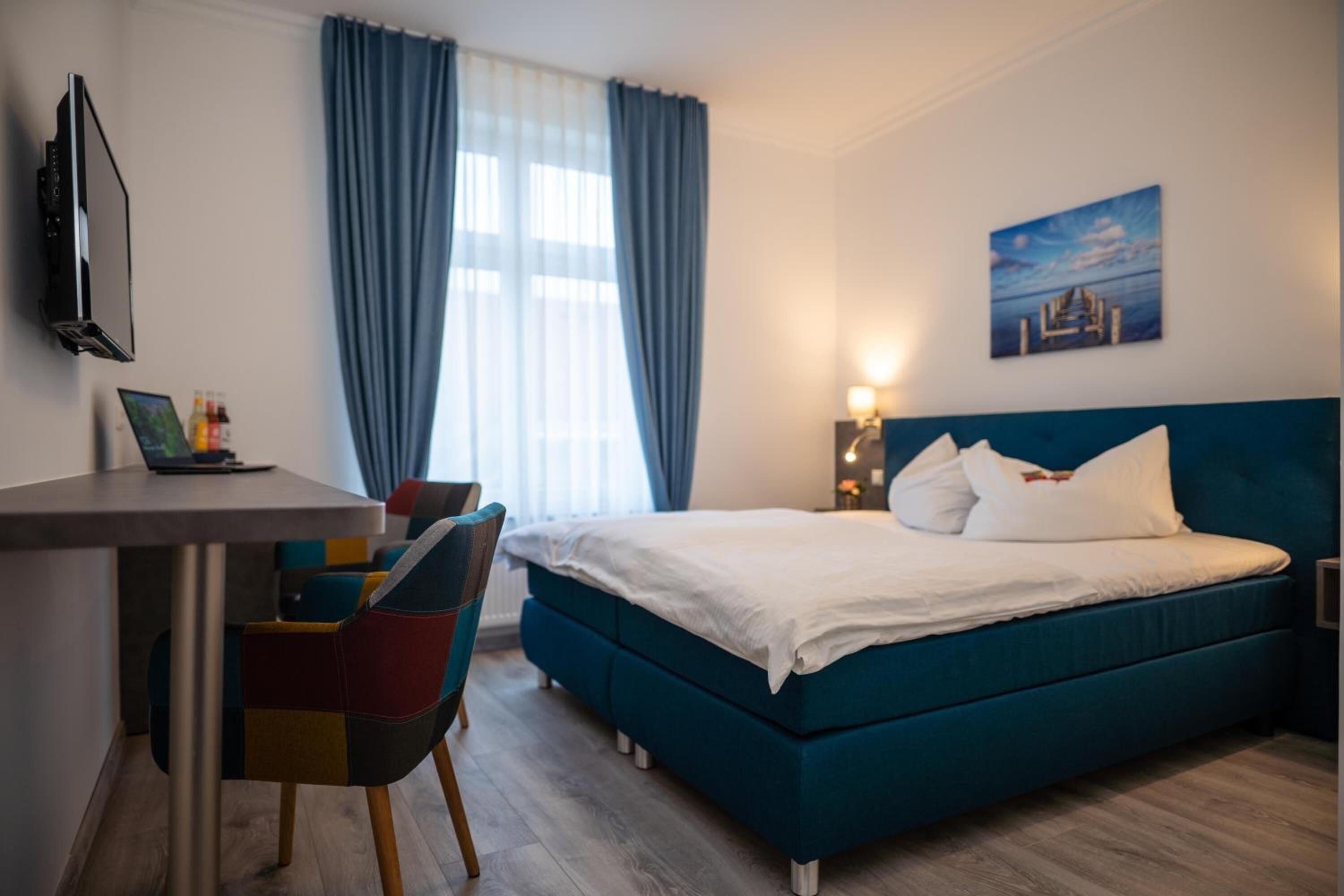 A modern hotel room with a teal bed, a desk with a laptop, colorful chairs, and a painting on the wall.