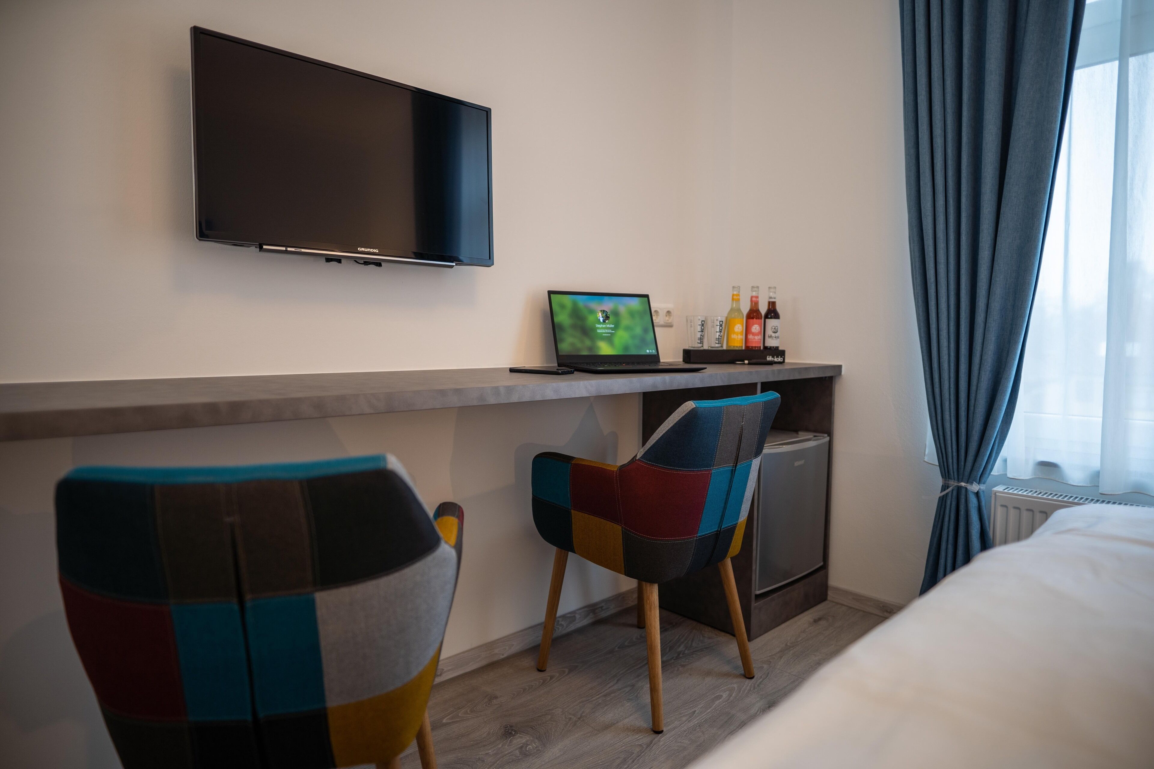 A hotel room with a flat-screen TV mounted on the wall, a laptop on a desk, two colorful chairs, a bed, and a window with curtains.