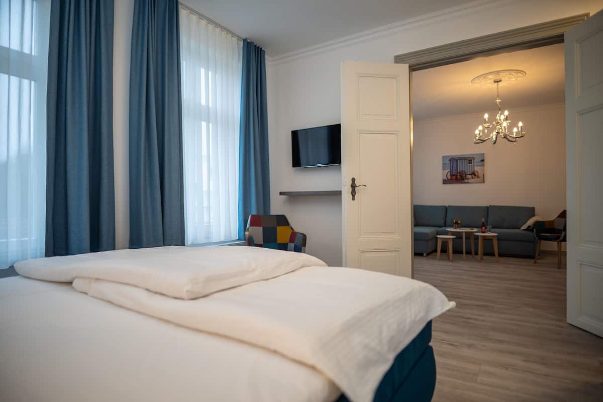 A hotel room with a large bed, a television mounted on the wall, a sofa set in the living area, and a chandelier.