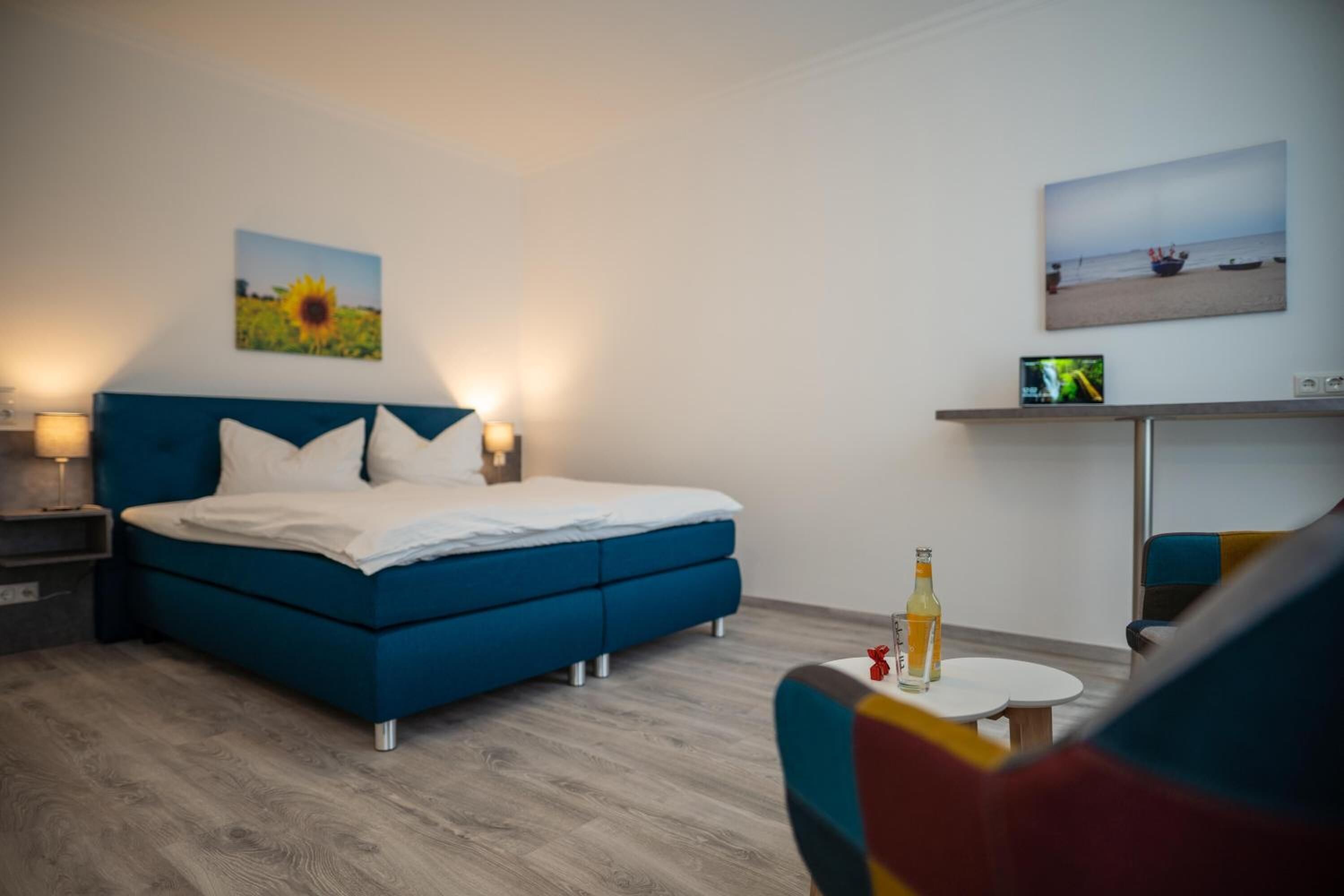 Ein modernes Schlafzimmer mit einem blauen Bett, weißen Kissen, einem Nachttisch, einem Fernseher auf einem Schrank und einem kleinen Tisch mit einer Flasche.