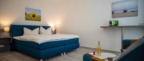 A modern bedroom with a blue bed, white pillows, a nightstand, a TV on a stand, and a small table with a bottle.