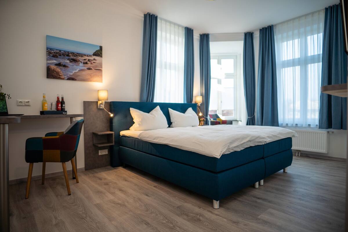 A modern hotel room with a large bed, a desk with a chair, and a view of the beach through the window.