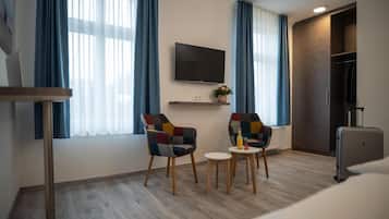 Una camera d'albergo moderna con una TV a schermo piatto montata a parete, due poltrone colorate, un piccolo tavolino con una bottiglia e una valigia sul pavimento.
