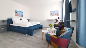 Une chambre d’hôtel moderne avec un grand lit, une petite table surmontée d’un vase de fleurs, un fauteuil coloré et un téléviseur fixé au mur.