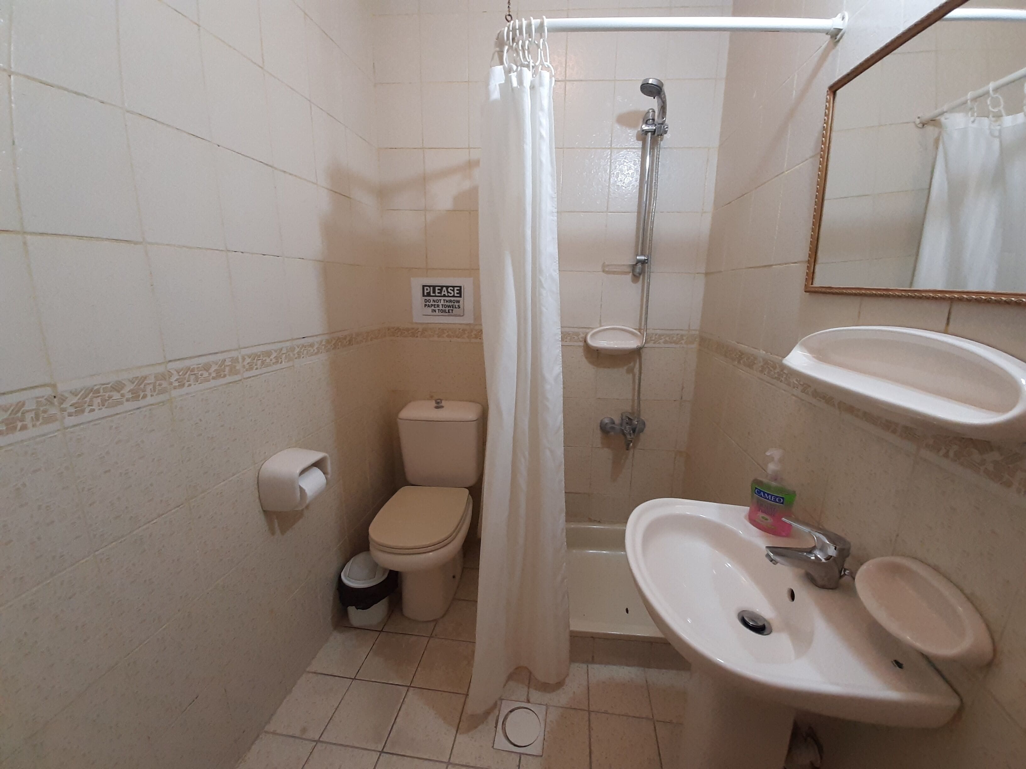Ein kleines Badezimmer mit Toilette, Dusche und Waschbecken. Ein Schild an der Wand weist darauf hin: "Bitte keine Papierhandtücher in die Toilette werfen.