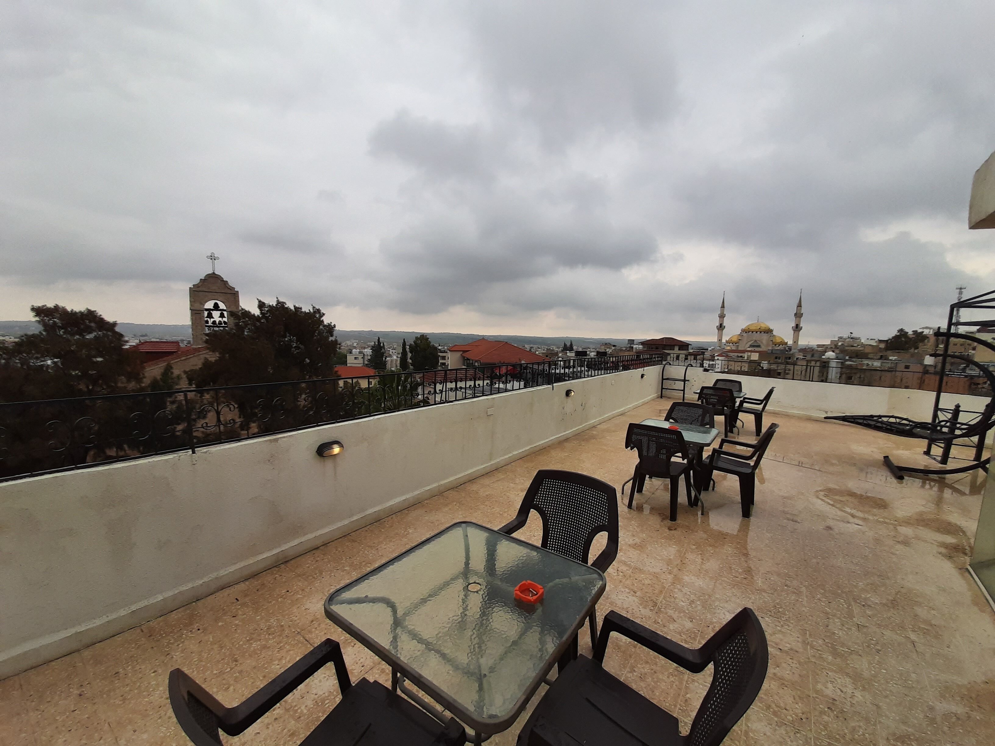 A rooftop terrace with a glass table and chairs, overlooking a cityscape with buildings and a mosque.