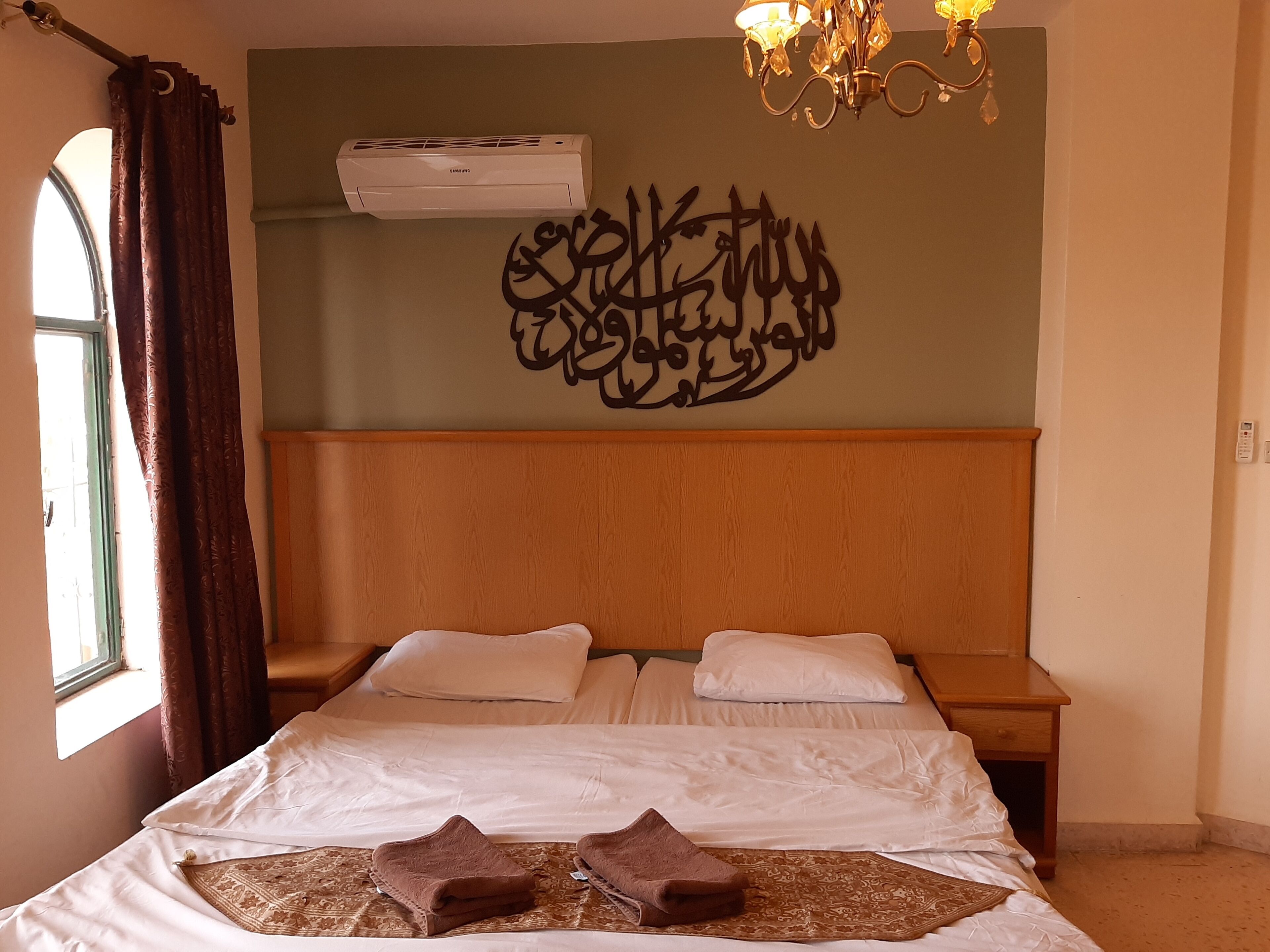 A bedroom with a bed, two pillows, a wooden headboard, a wall-mounted air conditioner, a chandelier, and a window with curtains.
