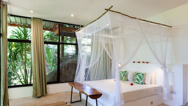A bedroom with a bed under a canopy, a bench, and a view of greenery outside. - Free Wifi + Outdoor shower at La Pointe Beach Huts (Praslin)