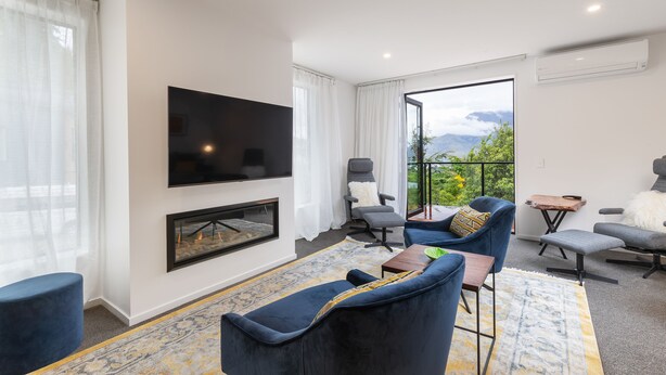 A modern living room with a flat-screen TV, comfortable seating, a fireplace, and a large window offering a scenic view. - MyHolidays - The Four Seasons Queenstown (4 brms + 4.5 bathrooms) (Queenstown)