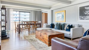 Ein geräumiges Wohnzimmer mit Couchtisch aus Holz, grauem Sofa und Ledersessel. Ein Essbereich mit Holztischen und Stühlen ist ebenfalls zu sehen.