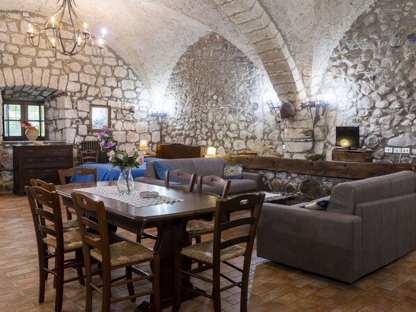 A stone-walled room with a dining table, chairs, sofas, and a television.