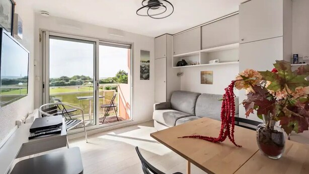 A modern living room with a sliding glass door leading to a balcony, a grey sofa, a wooden table, and a vase with flowers.