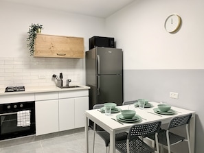 A modern kitchen with white cabinets, a black refrigerator, a wooden cabinet with a plant, and a dining table set with green plates and glasses.