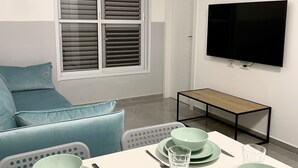 A modern living room with a sofa, dining table set with green plates and glasses, a TV mounted on the wall, and a window with blinds. - next to Soroka and BGU (Beer sheva)