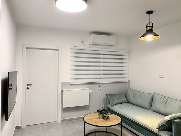 A modern living room with a sofa, a small round table with a plant, a TV, a ceiling light, and an air conditioning unit.
