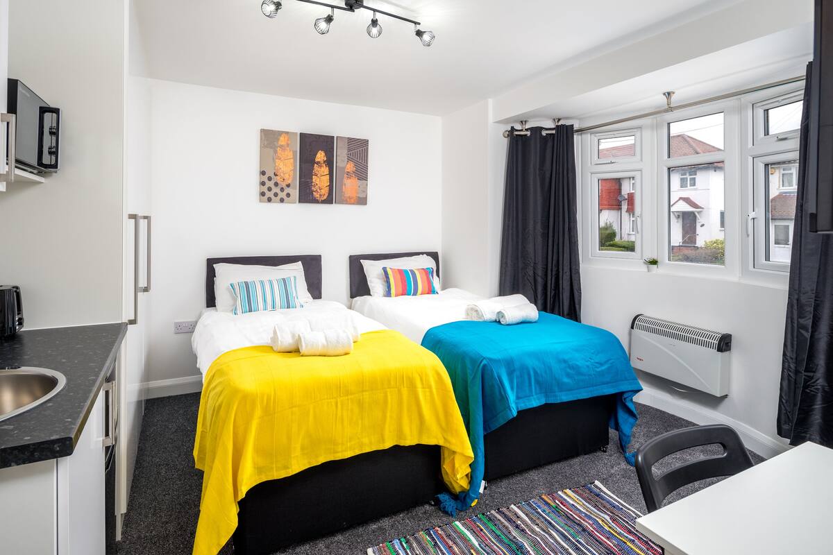 A hotel room with two beds, a kitchenette, a radiator, and a window with curtains.