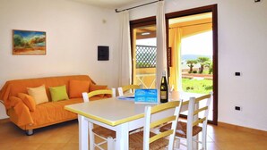 A dining area with a table and chairs, a sofa, a painting on the wall, and a view of the outdoors through an open door.
