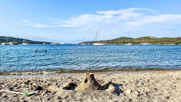 Una playa con arena en primer plano, mar tranquilo y veleros a lo lejos.