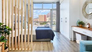 Et moderne hotelværelse med seng, skrivebord, stol, plante og udsigt over bybilledet gennem store vinduer.
