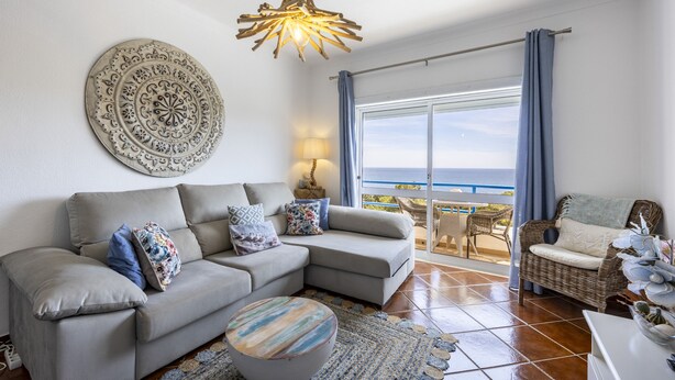 Una sala de estar con un sofá, una mesa redonda de centro, un adorno en la pared, una lámpara colgante y vista al mar a través de un ventanal.