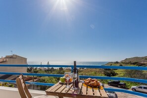 A wooden table with a bottle, glasses, and food on a balcony overlooking a coastal view.