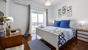 Habitación de hotel con cama, mesitas de noche, escritorio, silla, lámpara, un florero con flores, ventana con cortinas y una obra de arte en la pared.