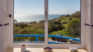 Una habitación con un ventanal grande que da a un paisaje costero, barandilla azul y una mesa con una taza y un plato.
