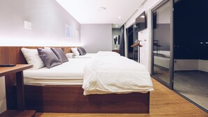 A modern hotel room with a large bed, a wooden bedside table, a flat-screen TV, and a sliding glass door leading to a balcony.