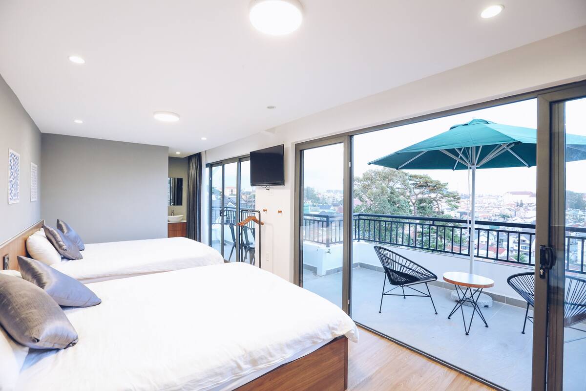Quarto de hotel com duas camas, uma varanda com mesa e cadeiras, e uma televisão fixada na parede.