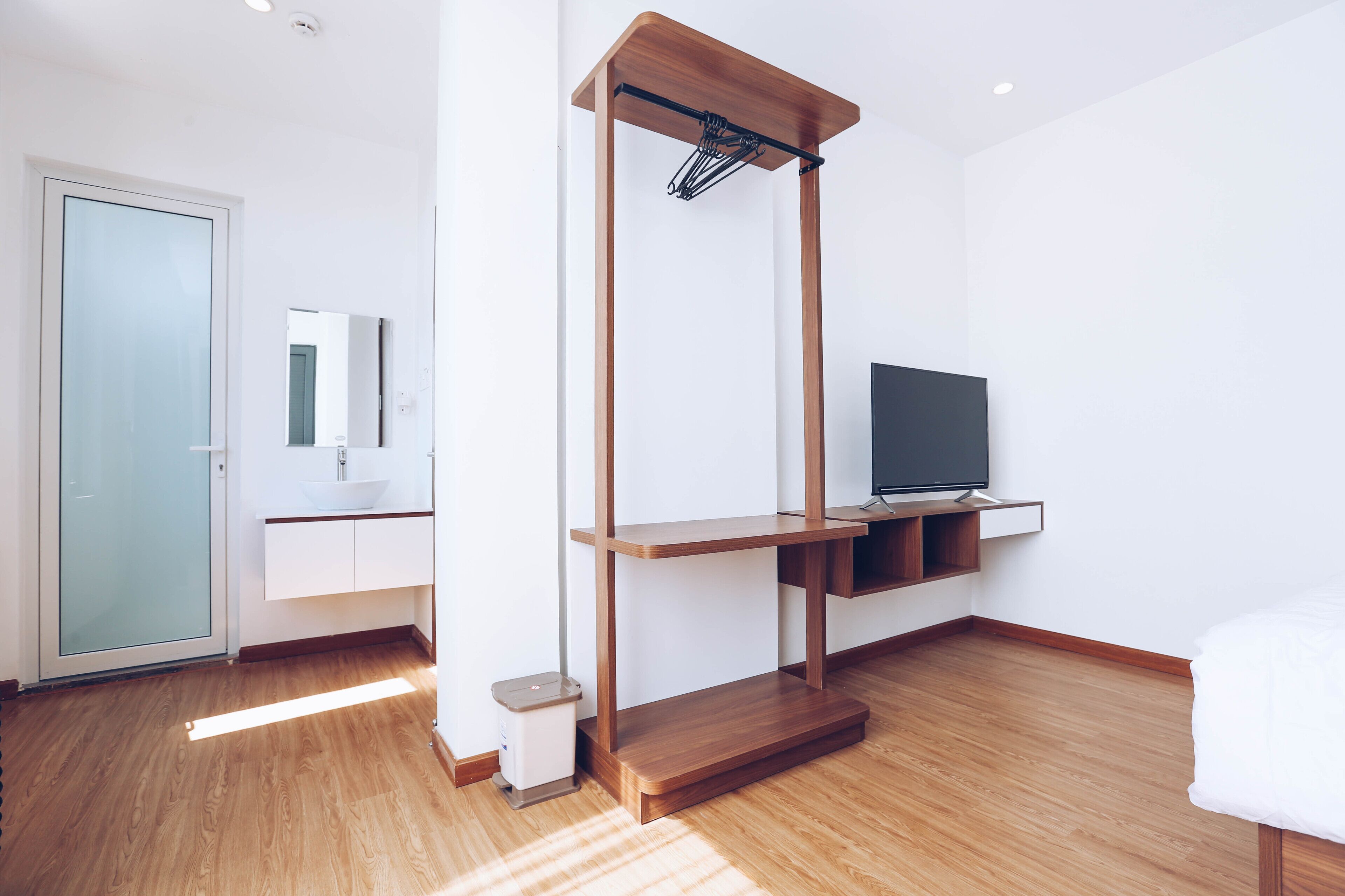 A modern room with a wooden floor, a TV on a shelf, a white bed, and a glass door.