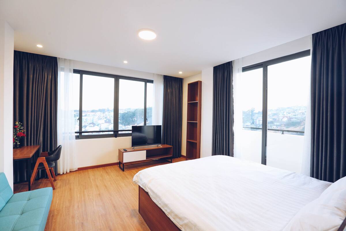 Quarto de hotel moderno com uma cama grande, televisão sobre um suporte de madeira, escrivaninha com cadeira e vista da cidade através de janelas amplas.