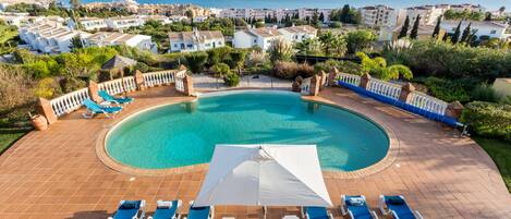A swimming pool with a surrounding patio area, lounge chairs, and a view of the sea and buildings in the background.