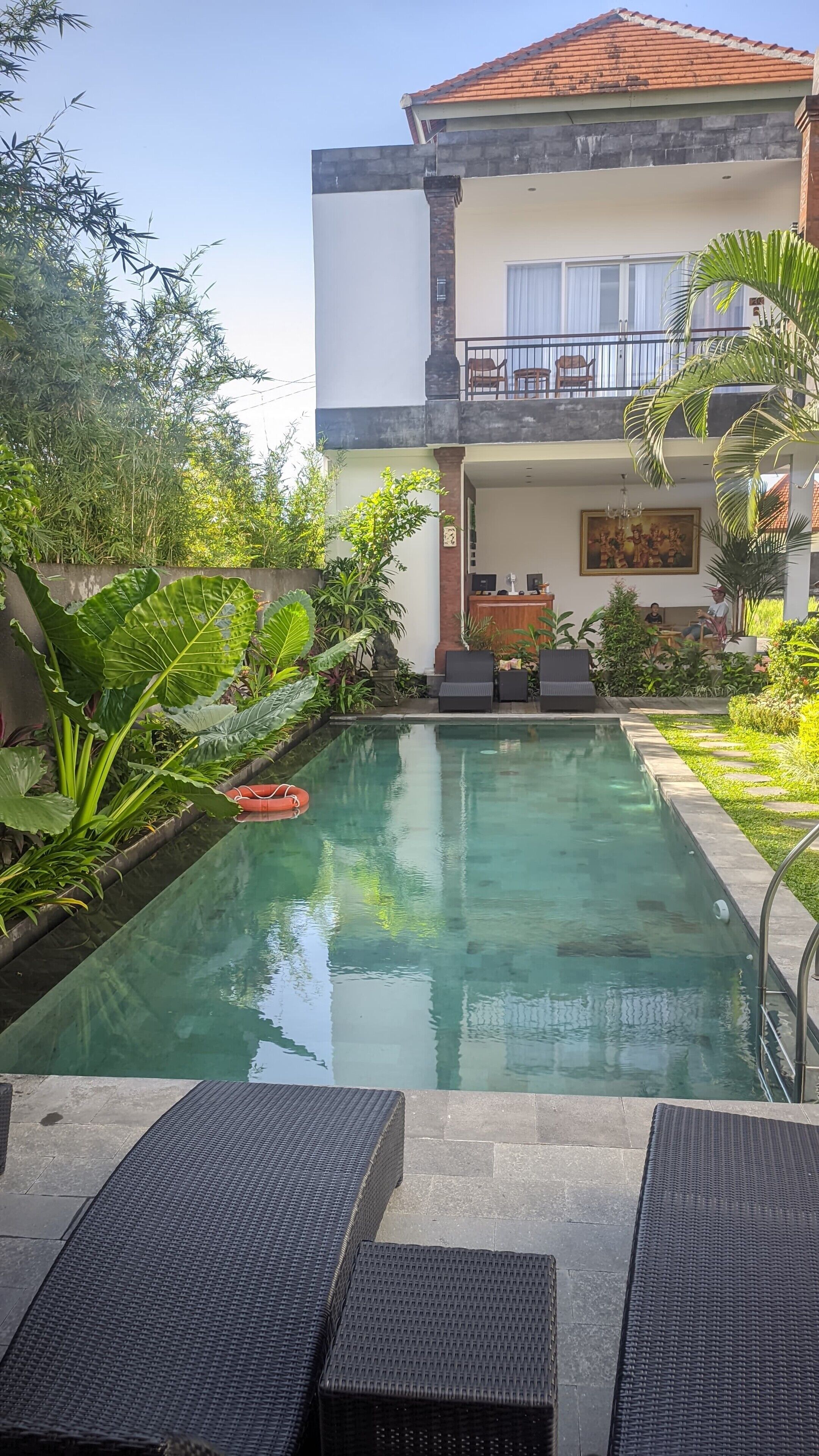 A swimming pool with lounge chairs, surrounded by lush greenery and a two-story building with a balcony.