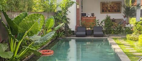 A swimming pool with lounge chairs, surrounded by lush greenery and a two-story building with a balcony.