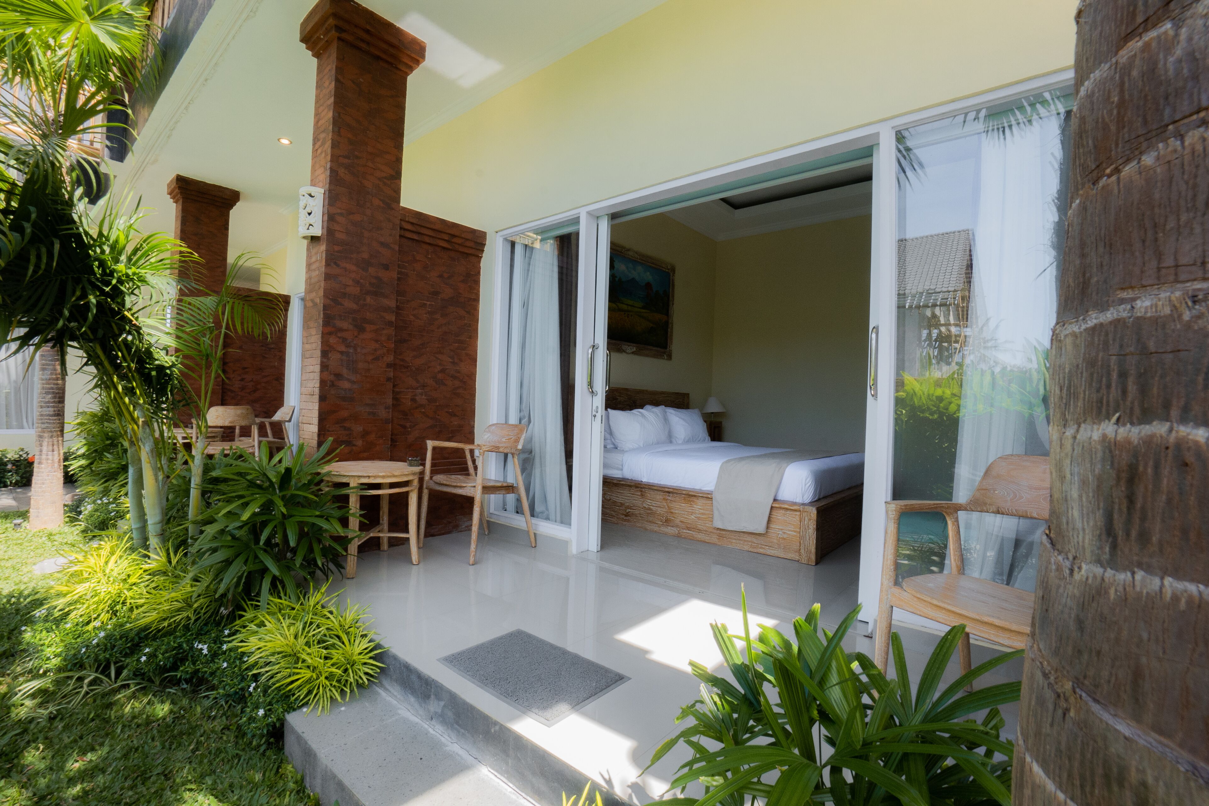 Una habitación de hotel moderna con un gran ventanal corredero que da a un balcón, una cama con cabecera blanca y una pequeña zona de estar con mesa y sillas.