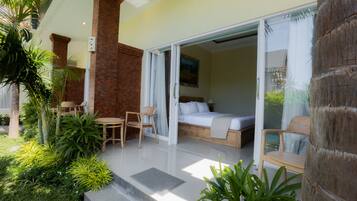 Una habitación de hotel moderna con un gran ventanal corredero que da a un balcón, una cama con cabecera blanca y una pequeña zona de estar con mesa y sillas.
