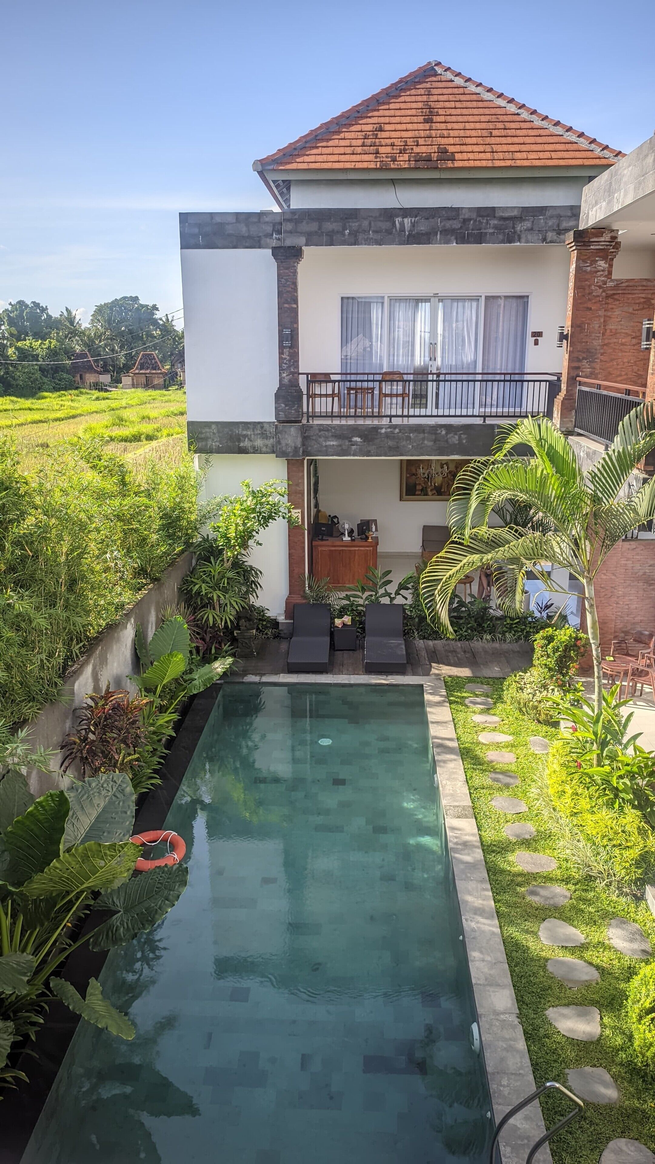 A house with a swimming pool, a balcony with chairs, and lush greenery.