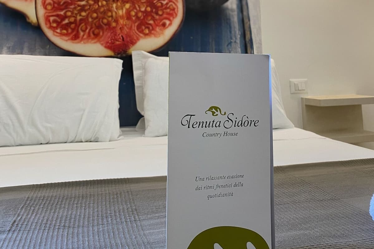 A hotel room with a large poster of figs, a white bed, and a sign reading "Tenuta Sidore Country House".