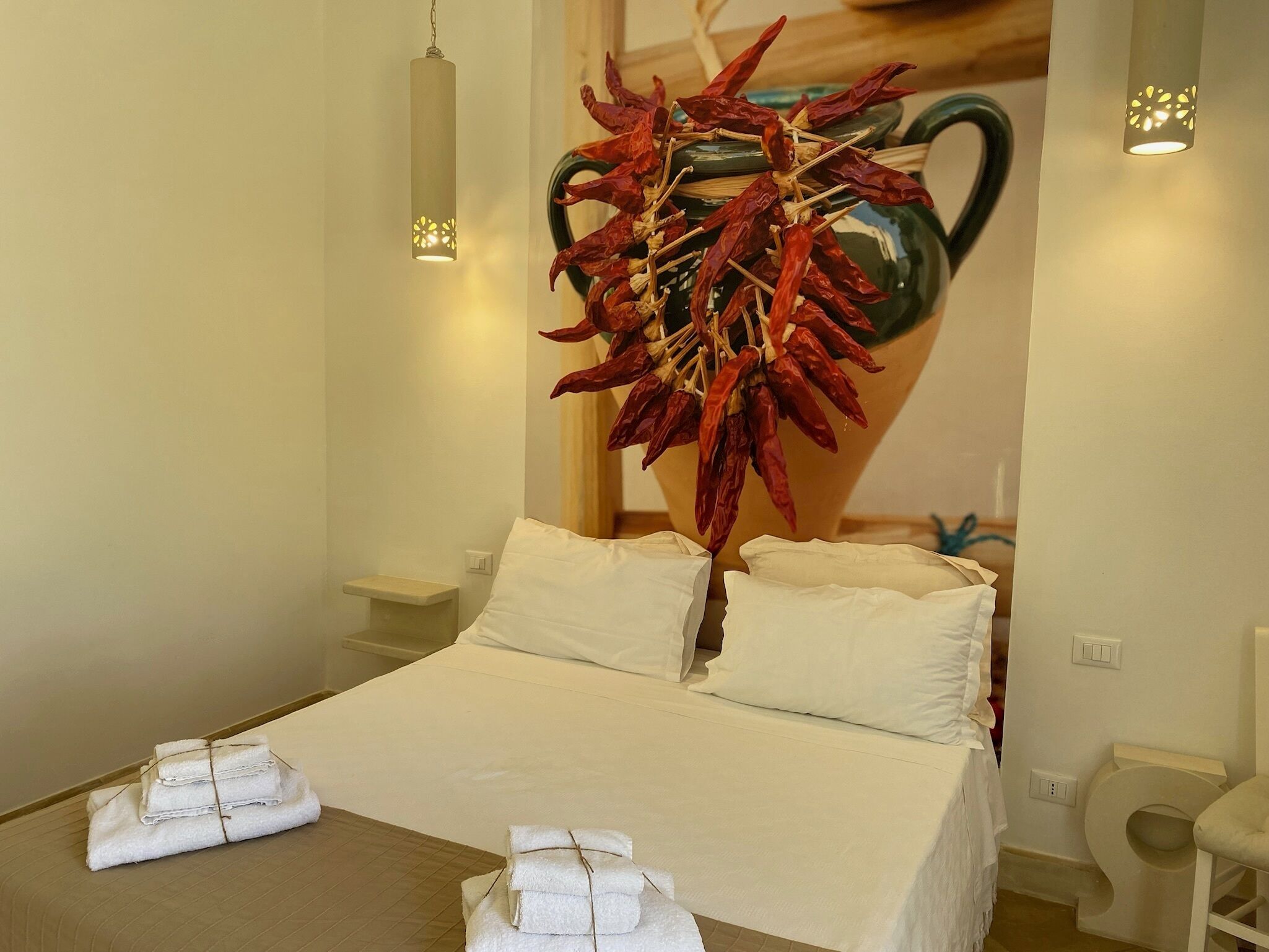 Ein Hotelzimmer mit einem Bett, zwei Nachttischen, einer an der Wand befestigten Vase mit getrockneten roten Chilischoten und zwei Wandleuchten.