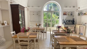 A bright, airy kitchen with white cabinetry, wooden tables, and chairs. There are potted plants adding a touch of greenery. - Tenuta Sidore (Cursi)