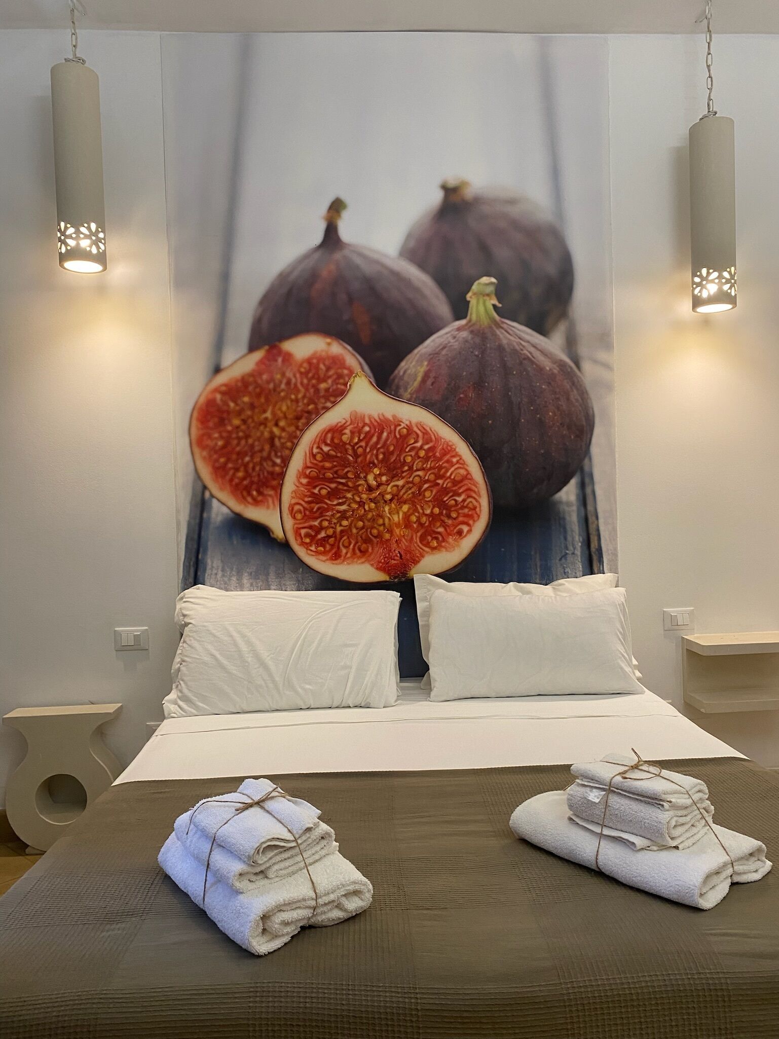 A hotel room with a large bed, white pillows, and towels. There is a wall art of figs and a light fixture on the ceiling.
