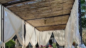 An outdoor patio with a wooden table and white chairs, covered by a bamboo canopy and draped with white fabric. - Tenuta Sidore (Cursi)