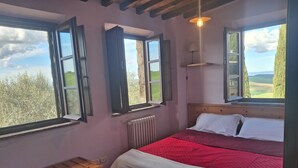 A bedroom with a bed, a wooden bench, a radiator, and large windows with open shutters. - House with private garden in the Crete Senesi (Asciano)