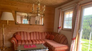 Una sala de estar acogedora con suelo de madera, un sofá a rayas, una mesa de centro de vidrio, una araña, un espejo y una ventana con cortinas.