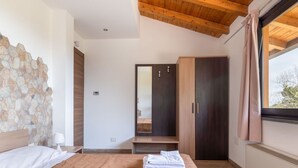Une chambre avec un mur en pierre, un plafond en bois, un lit recouvert d’une couverture marron, une armoire en bois, un miroir et une fenêtre avec des rideaux.