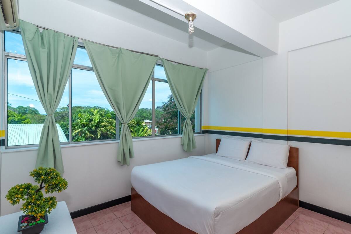 A hotel room with a bed, a window with curtains, a small table with a plant, and a view of greenery outside.