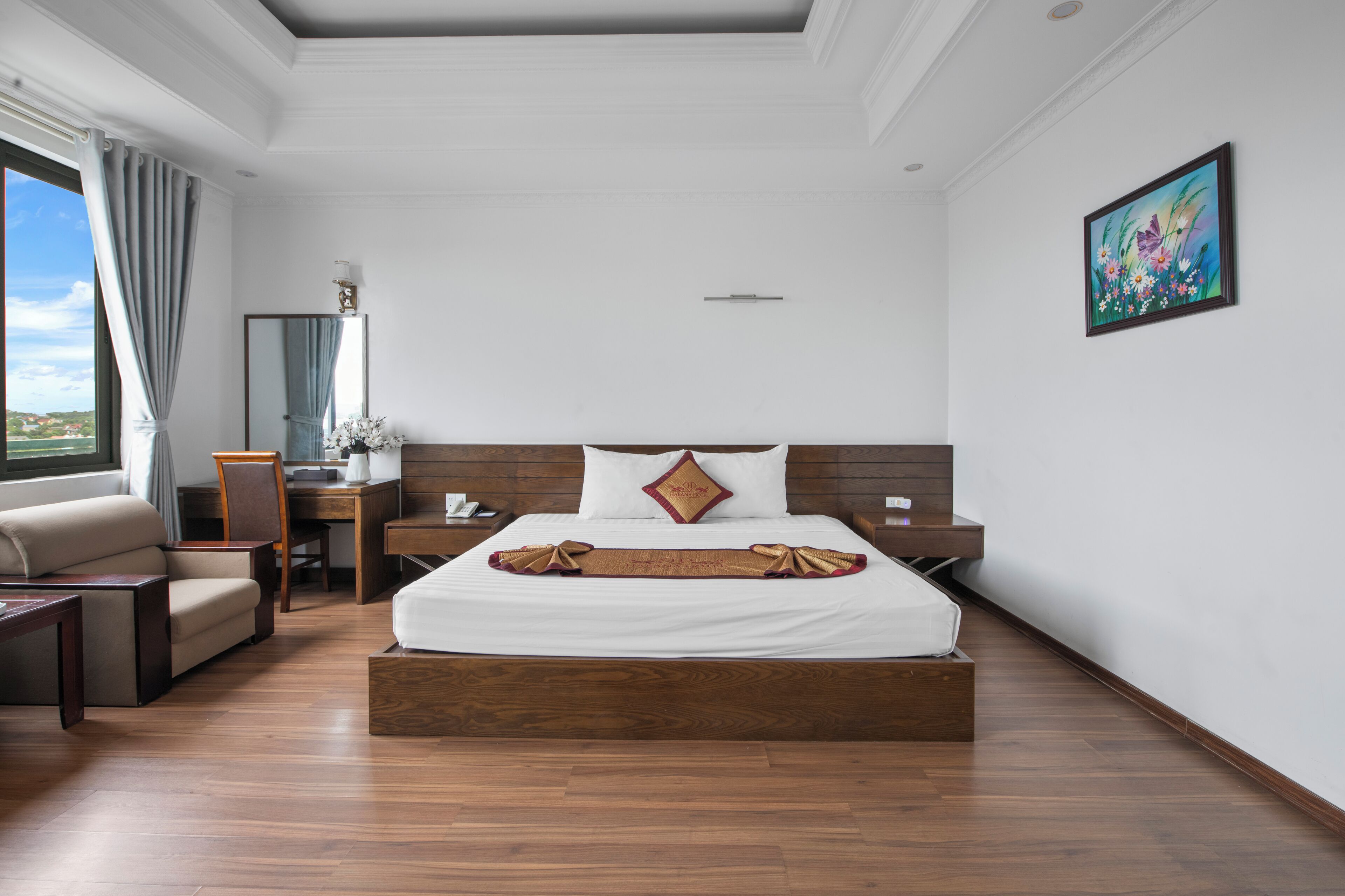 Habitación de hotel con una cama grande, cabecero de madera, un escritorio con silla, un sofá y una ventana con cortinas.
